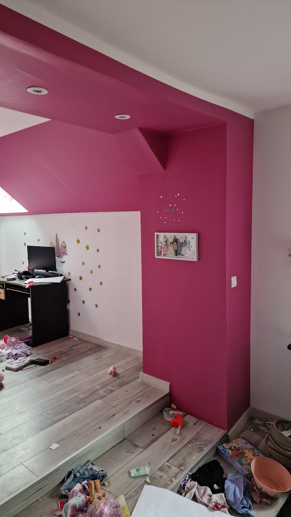 Pokój po malowaniu ścian i sufitu na intensywny róż, widoczne drewniane panele podłogowe, białe ściany z dekoracjami i meble. Stan przed sprzątaniem.