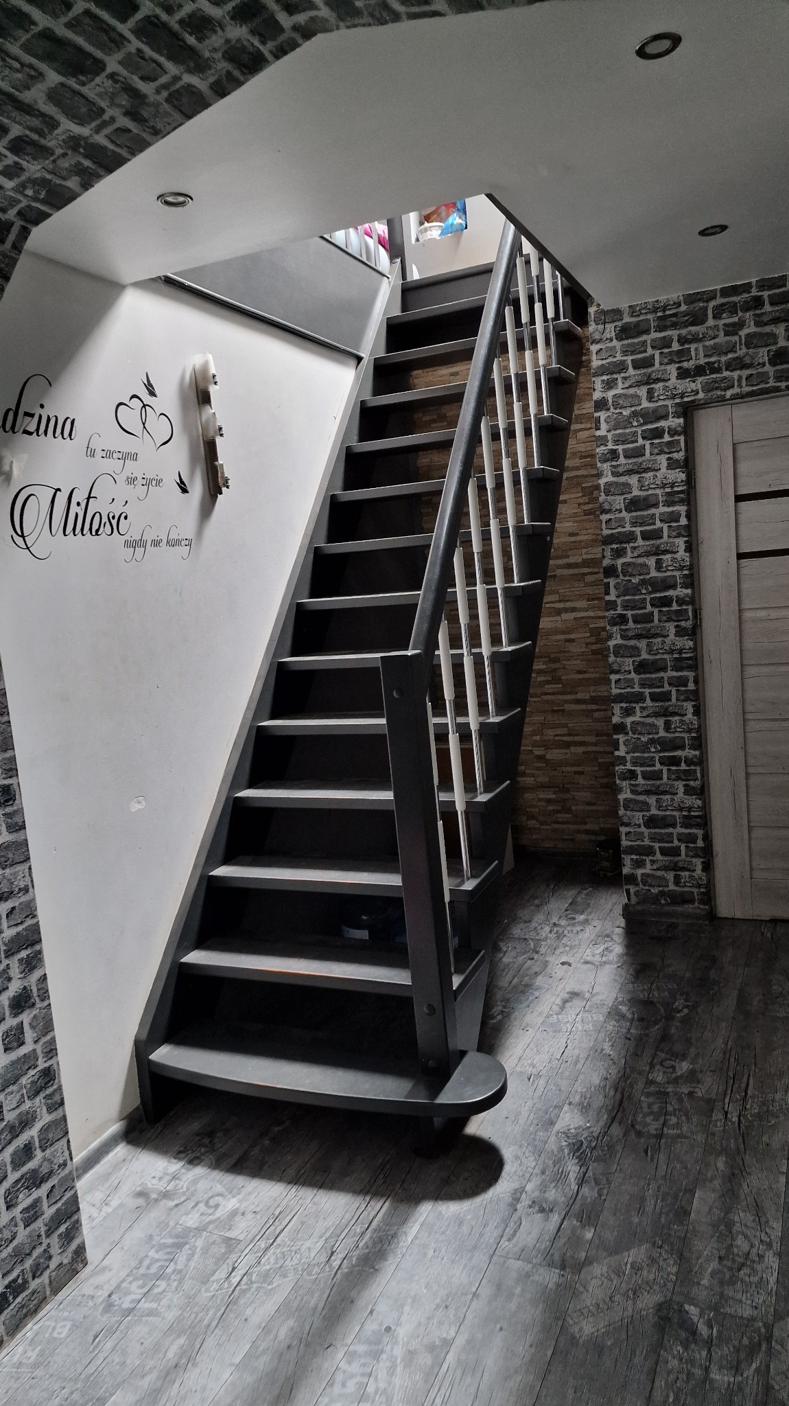 Schody w ciemnej tonacji z białymi balustradami, ściana z dekoracyjnym napisem, imitacja cegły na ścianach i podłodze, oświetlenie punktowe w suficie.