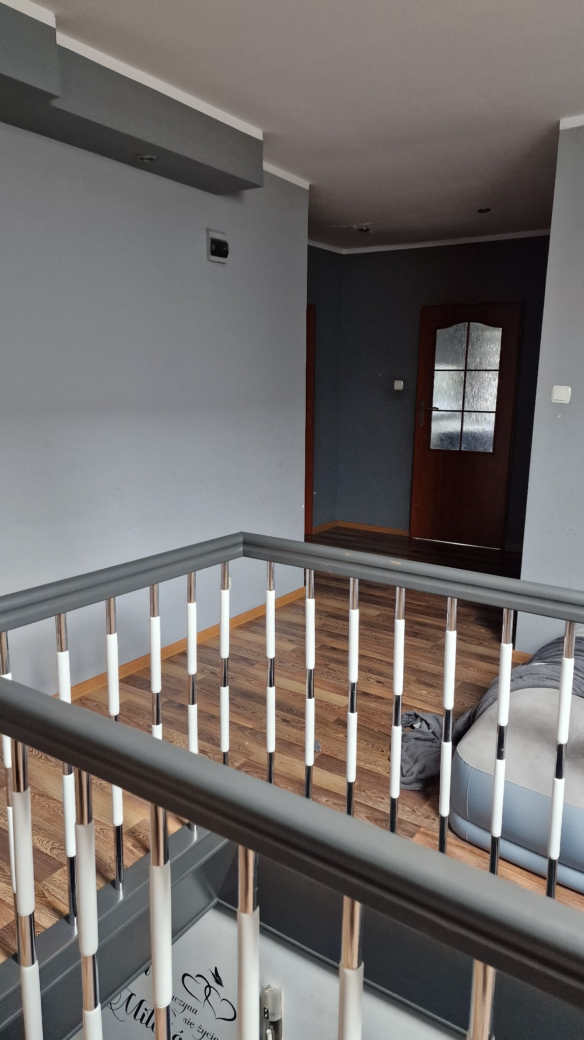 Szare ściany i drewniana podłoga widoczne zza szarej balustrady z białymi elementami. W tle brązowe drzwi z szybą i szara ściana.