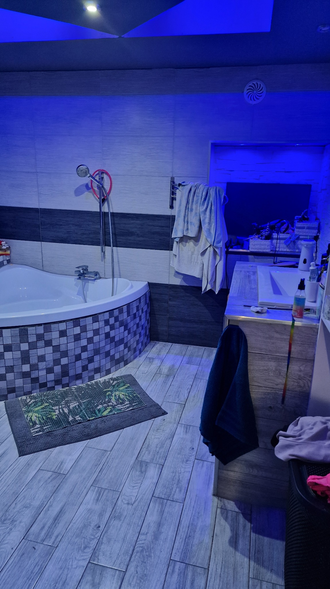 Nowoczesna łazienka z wanną narożną, wyłożona płytkami w odcieniach szarości i bieli. Uwagę przyciąga oświetlenie LED w kolorze niebieskim, nadające wnętrzu unikalny charakter.