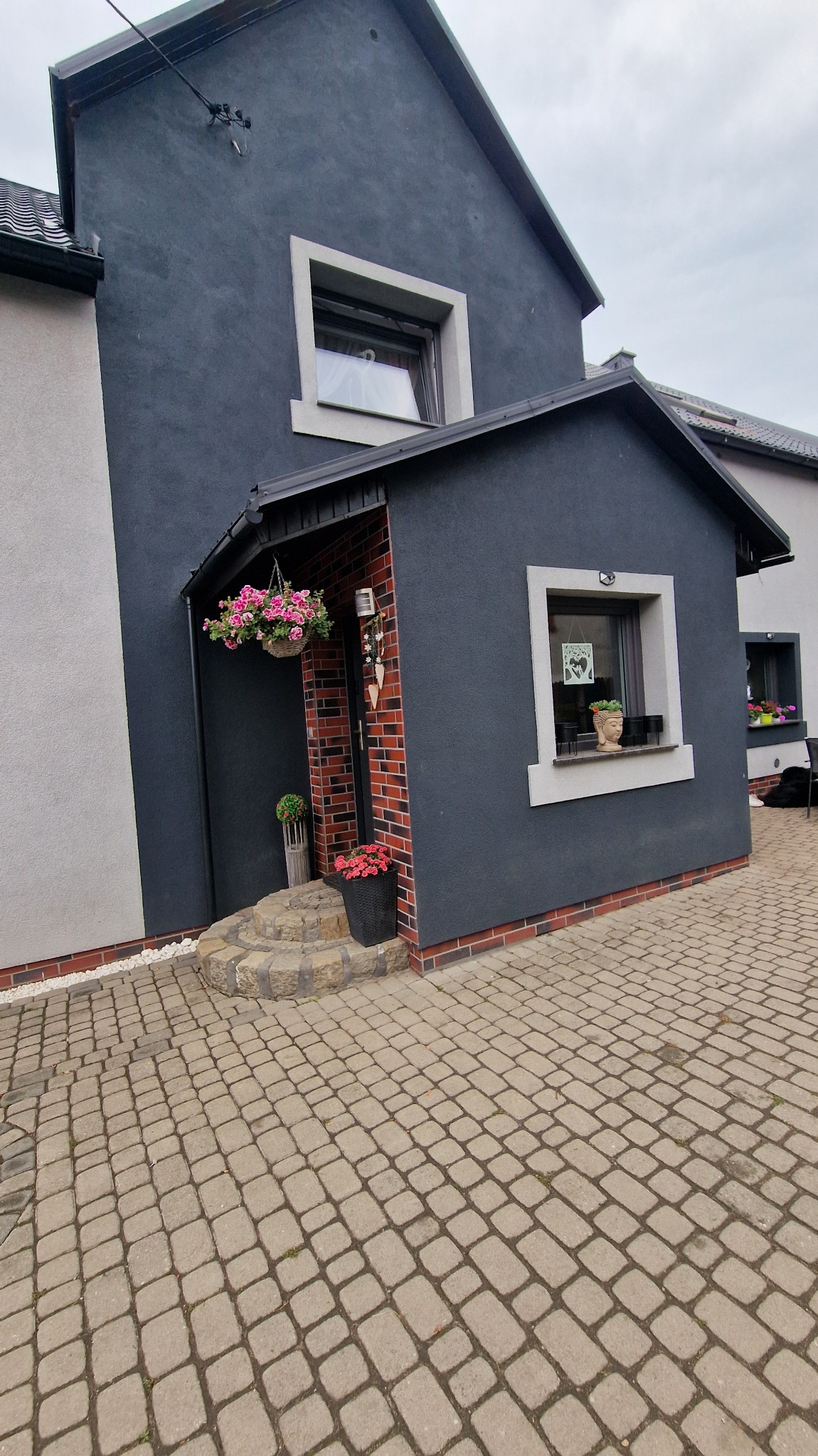 Elewacja domu z ciemnoszarą strukturą, ceglanym obramowaniem i jasnymi oknami. Wejście z doniczkami i wiszącymi kwiatami. Brukowany podjazd.