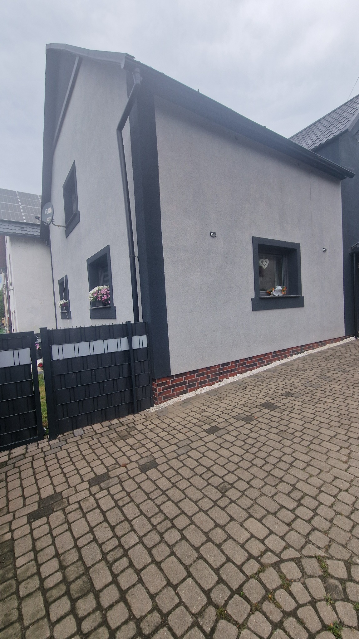 Elewacja domu z szarą strukturą i ciemnymi obramowaniami okien, z widoczną kostką brukową i fragmentem ogrodzenia. W oknach kwiaty i dekoracje.