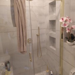 Beata Kraska - Nowoczesna kabina prysznicowa z marmurowymi płytkami, złotymi detalami i wnękami w ścianie. Na szklanej ściance wisi beżowy ręcznik, a obok stoi doniczka z orchideami.