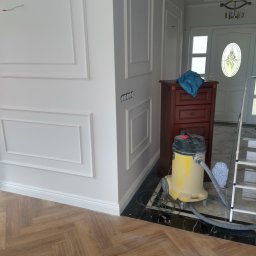Beata Kraska - Wykończone wnętrze z dekoracyjnymi listwami ściennymi, drewnianą podłogą w jodełkę i widocznym odkurzaczem. Widoczny fragment drzwi wejściowych.