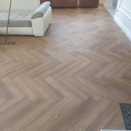 Beata Kraska - Ukończona podłoga z paneli ułożonych w jodełkę, widok z perspektywy. W tle meble i sofa. Podłoga w odcieniach brązu, jasne ściany.