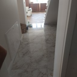 Beata Kraska - Wykończone marmurowe płytki podłogowe w korytarzu z widokiem na schody i salon. Widoczna skrzynka rewizyjna na ścianie i częściowo widoczne drzwi.