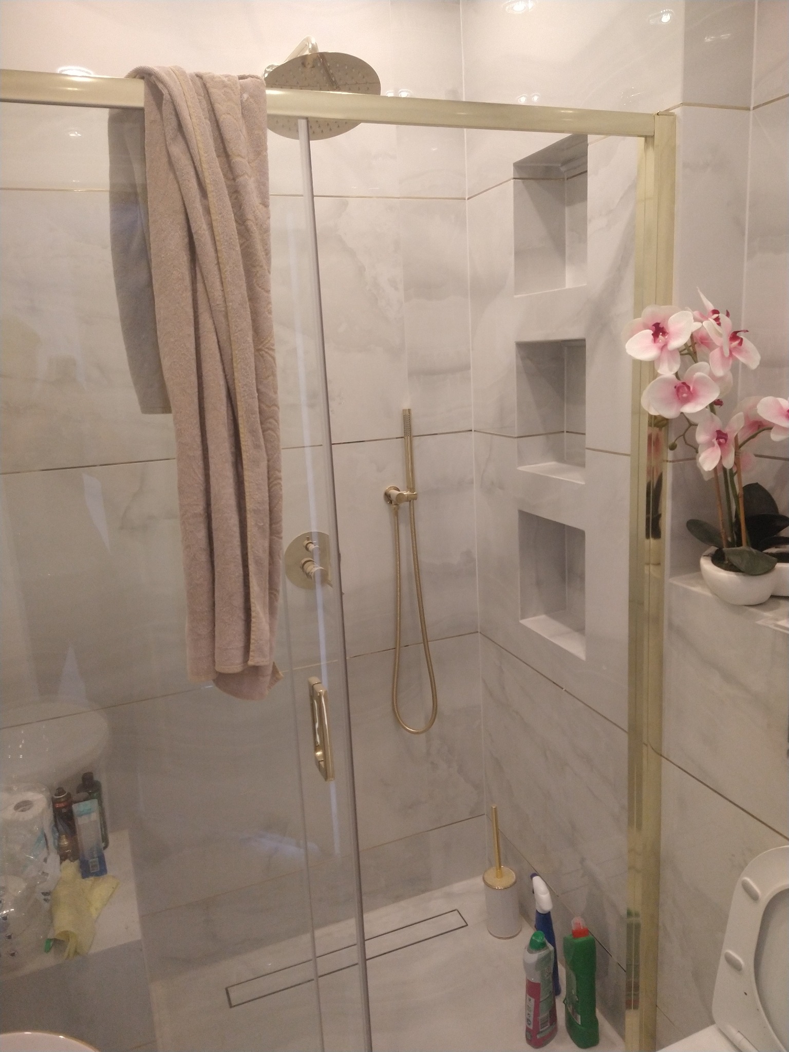 Nowoczesna kabina prysznicowa z marmurowymi płytkami, złotymi detalami i wnękami w ścianie. Na szklanej ściance wisi beżowy ręcznik, a obok stoi doniczka z orchideami.