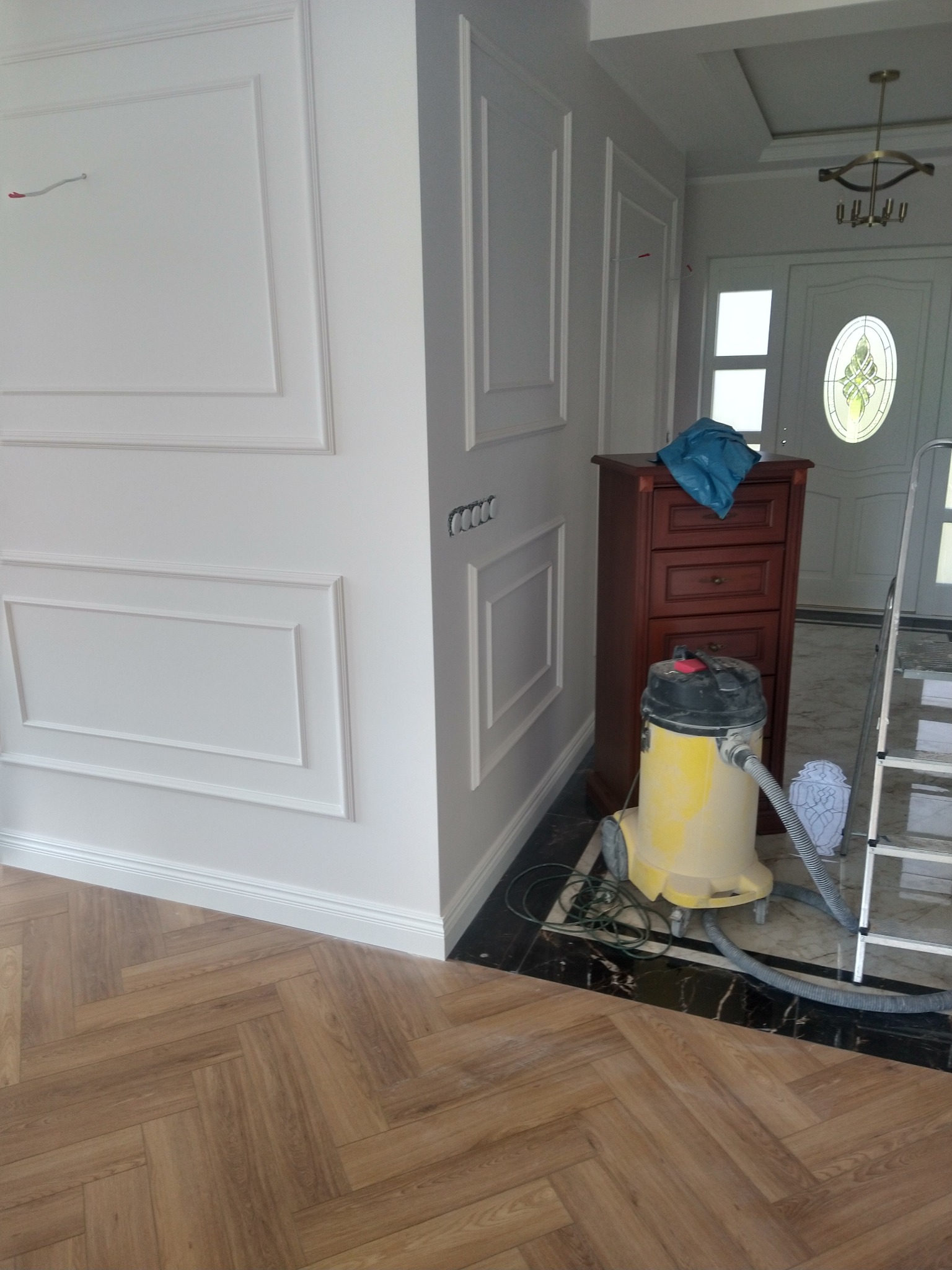 Wykończone wnętrze z dekoracyjnymi listwami ściennymi, drewnianą podłogą w jodełkę i widocznym odkurzaczem. Widoczny fragment drzwi wejściowych.