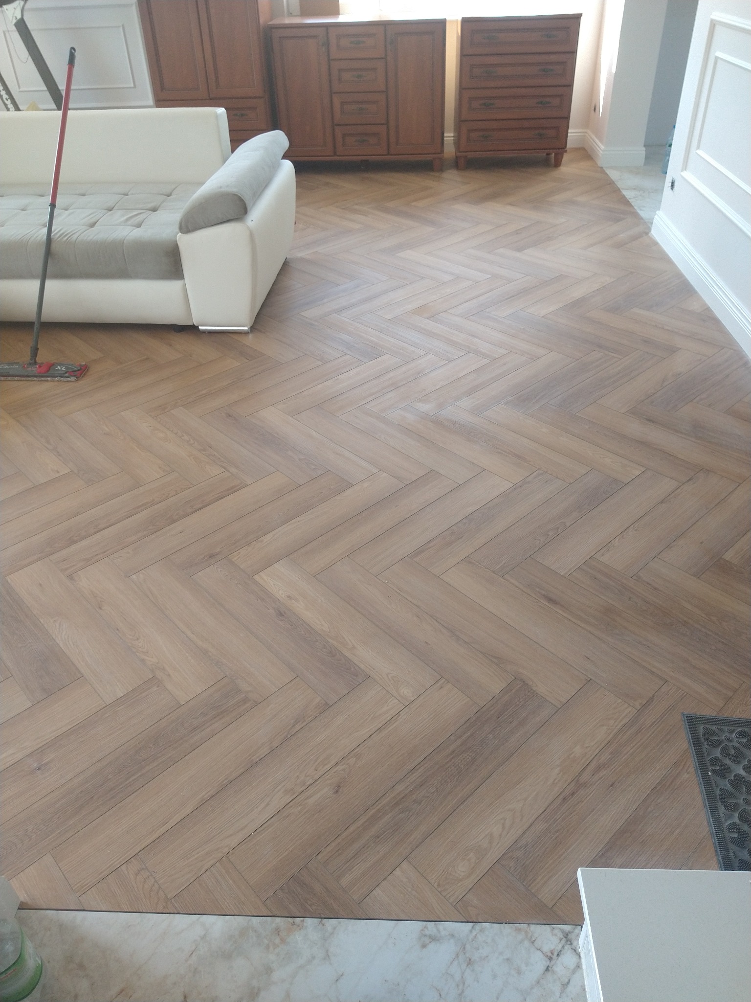 Ukończona podłoga z paneli ułożonych w jodełkę, widok z perspektywy. W tle meble i sofa. Podłoga w odcieniach brązu, jasne ściany.