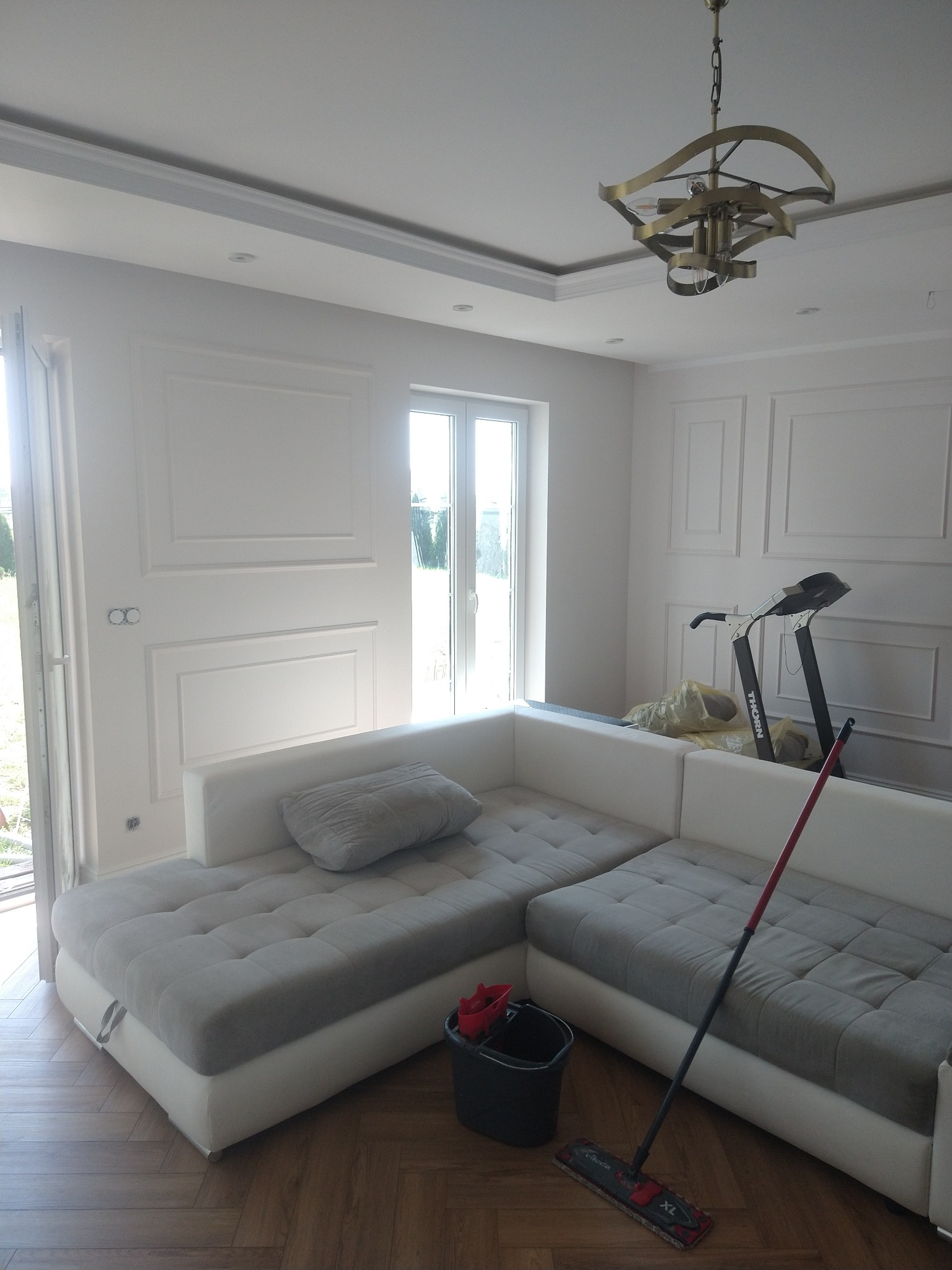 Białe, eleganckie wnętrze po remoncie: sofa narożna, parkiet w jodełkę, dekoracyjne panele ścienne. Widoczny mop i wiadro, sugerujące świeże sprzątanie po pracach wykończeniowych.