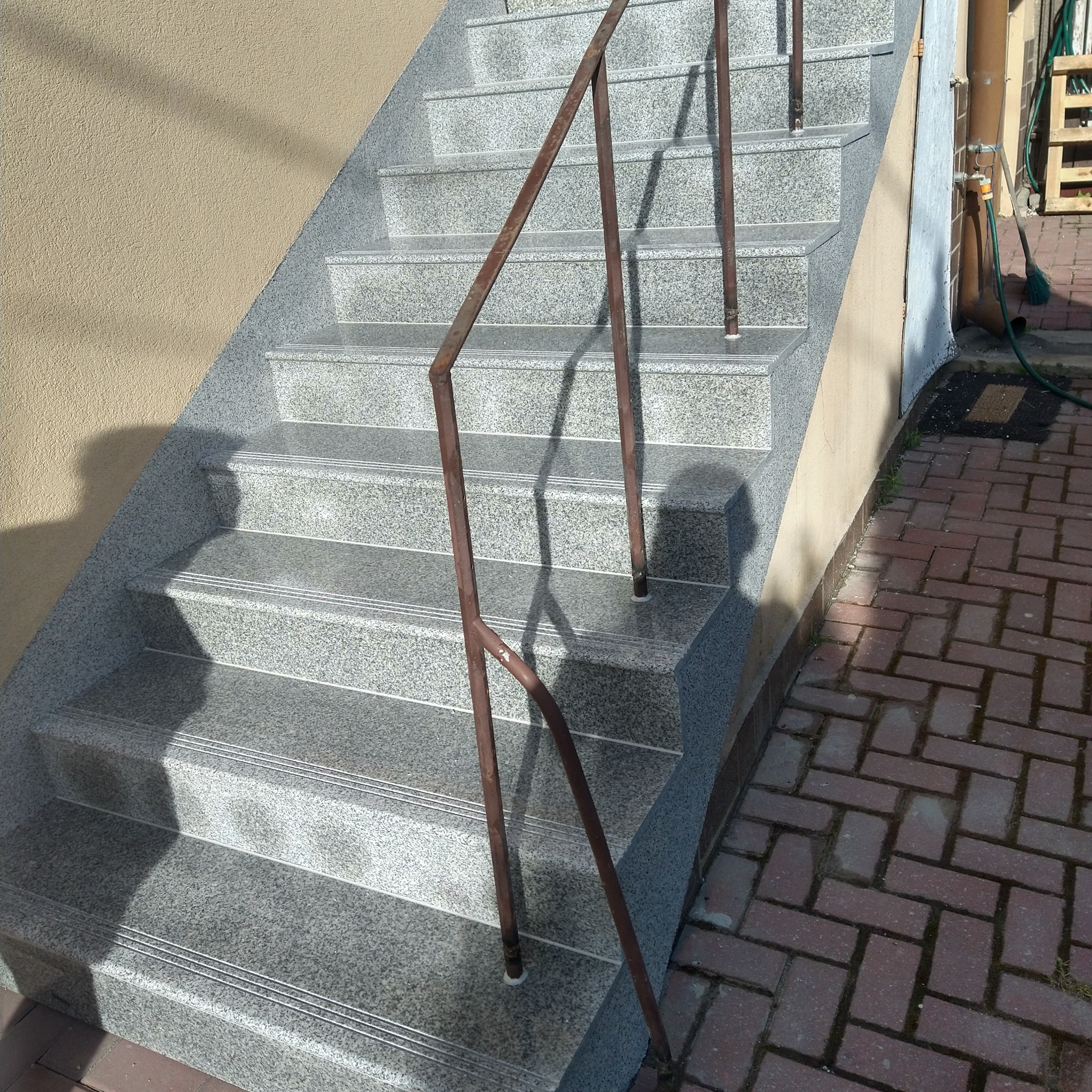 Zewnętrzne schody z szarego granitu z metalową, rdzawą barierką. Widoczne detale wykończenia stopni i faktura kamienia. Obok ściana budynku i chodnik z kostki.