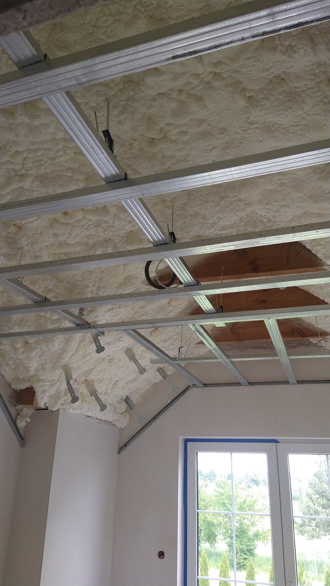 Izolacja pianą PUR poddasza z widoczną konstrukcją metalową sufitu podwieszanego i oknem. Detal wykończenia wnętrza w trakcie prac.