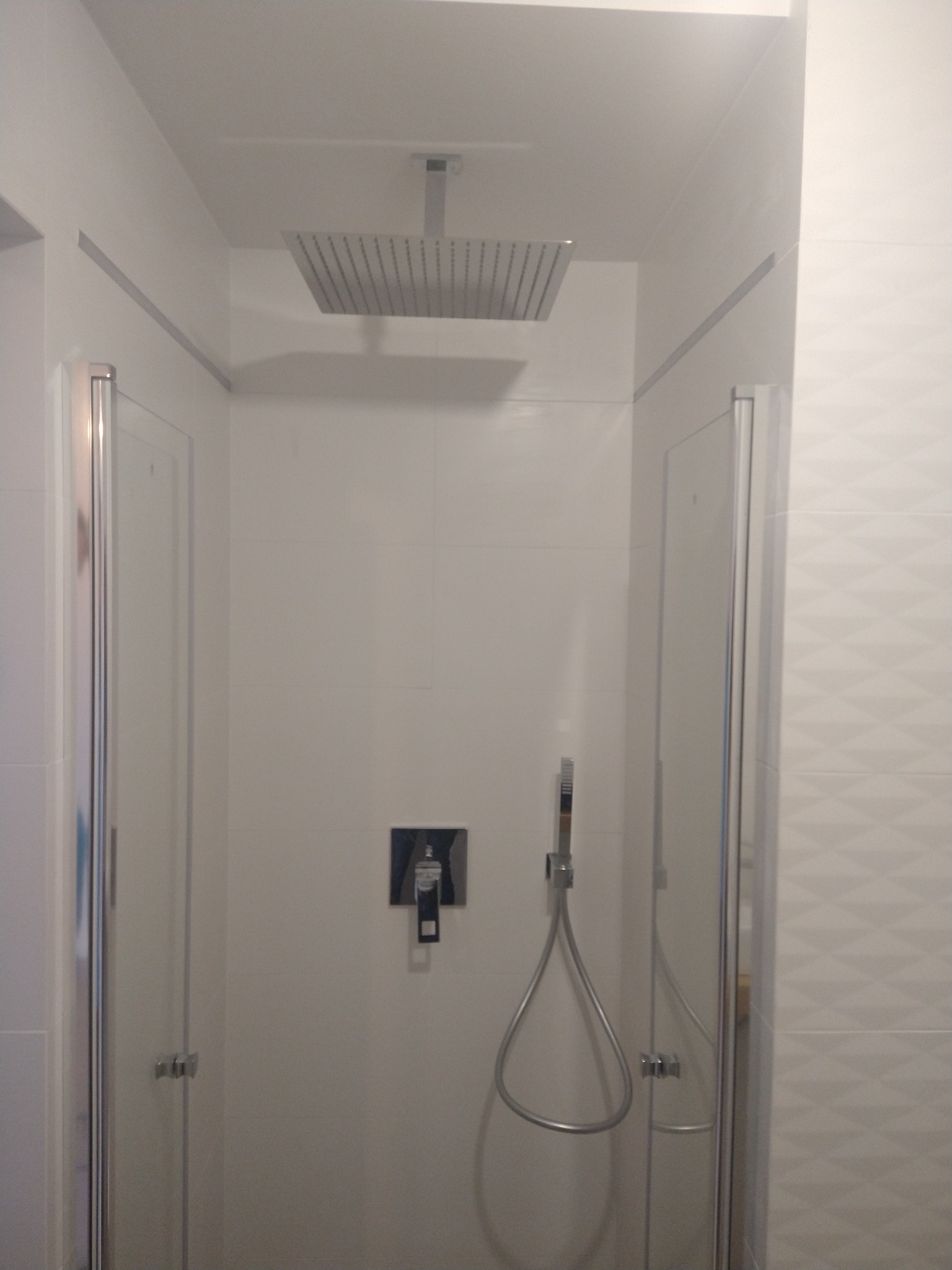 Nowoczesna kabina prysznicowa z deszczownicą i szklanymi drzwiami, wykończona białymi płytkami. Minimalistyczny design, chromowane elementy, widoczny panel sterowania.