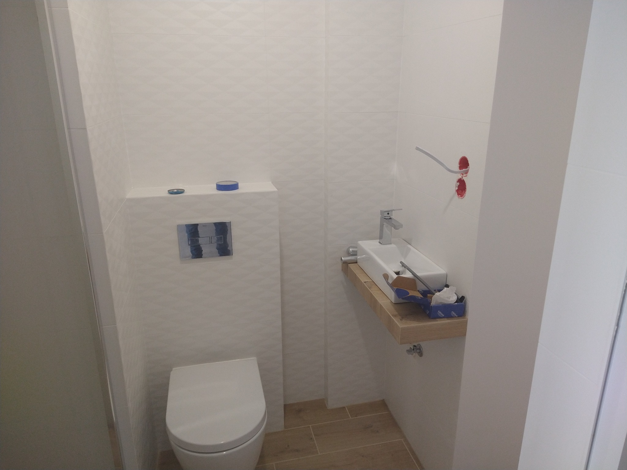 Nowoczesna, jasna toaleta z podwieszaną miską WC i umywalką na drewnianym blacie. Białe płytki z subtelnym wzorem, widoczne elementy instalacji elektrycznej.