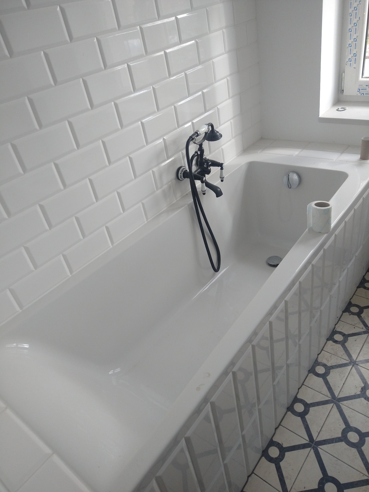 Biała wanna z czarną armaturą i białą glazurą typu metro w łazience. Na wannie rolka papieru toaletowego. Podłoga wyłożona płytkami w geometryczny wzór.