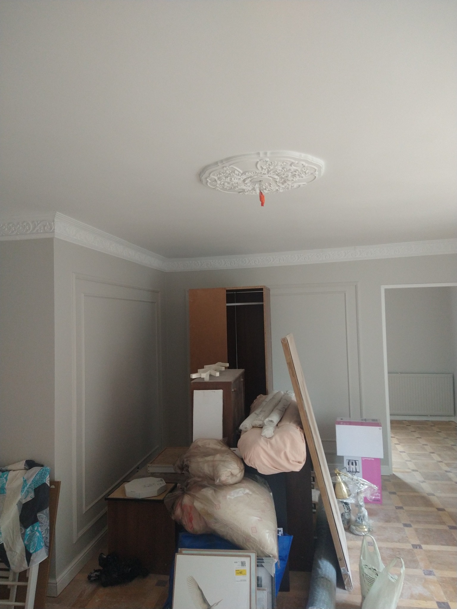 Pokój w trakcie wykończenia: ściany z dekoracyjnymi panelami, sztukateria sufitowa z rozetą. Część mebli i materiałów budowlanych zgromadzona w pomieszczeniu.