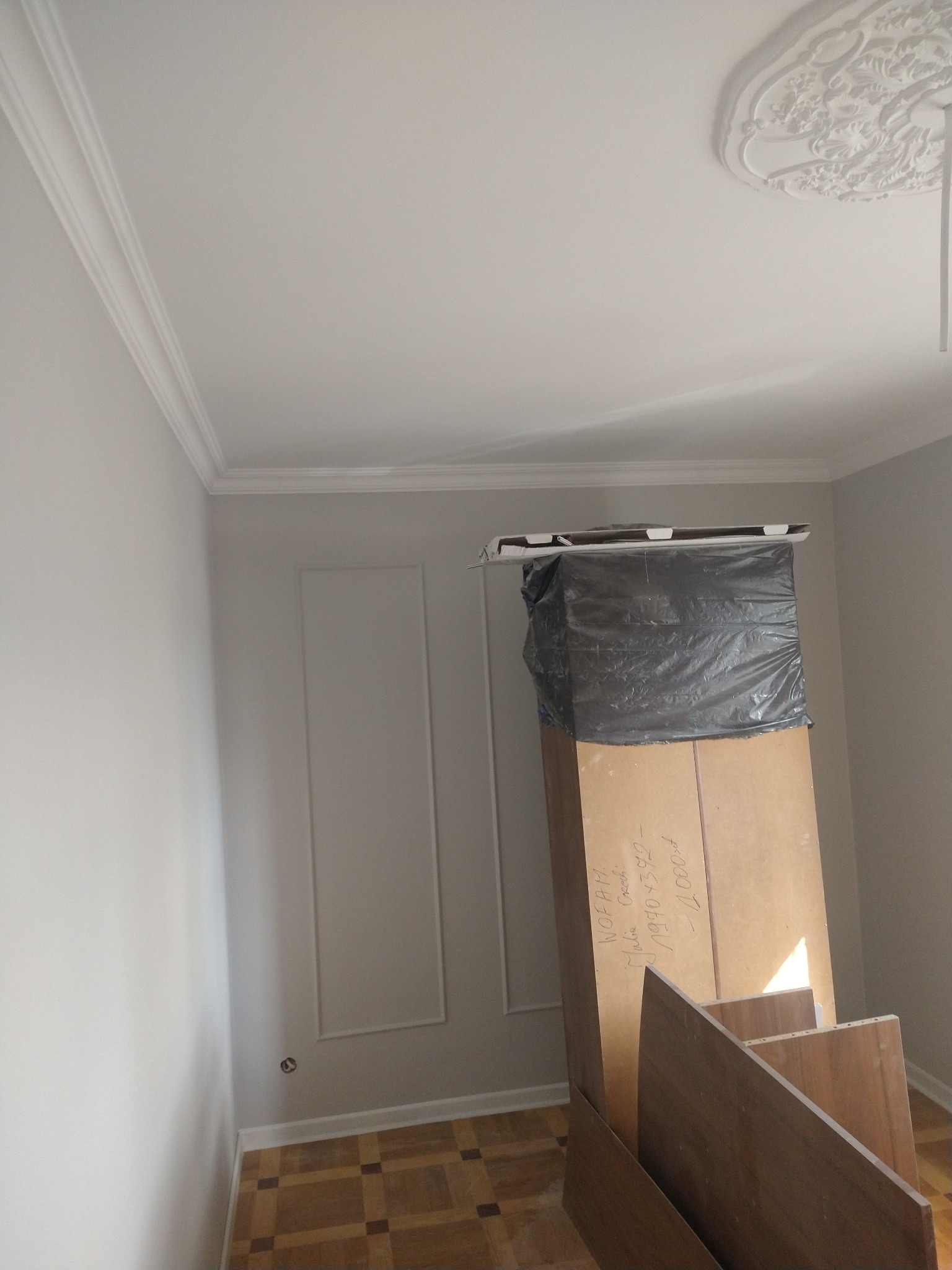 Białe ściany z dekoracyjną sztukaterią i listwami przypodłogowymi. Na podłodze parkiet. W rogu pokoju elementy mebli zabezpieczone folią.