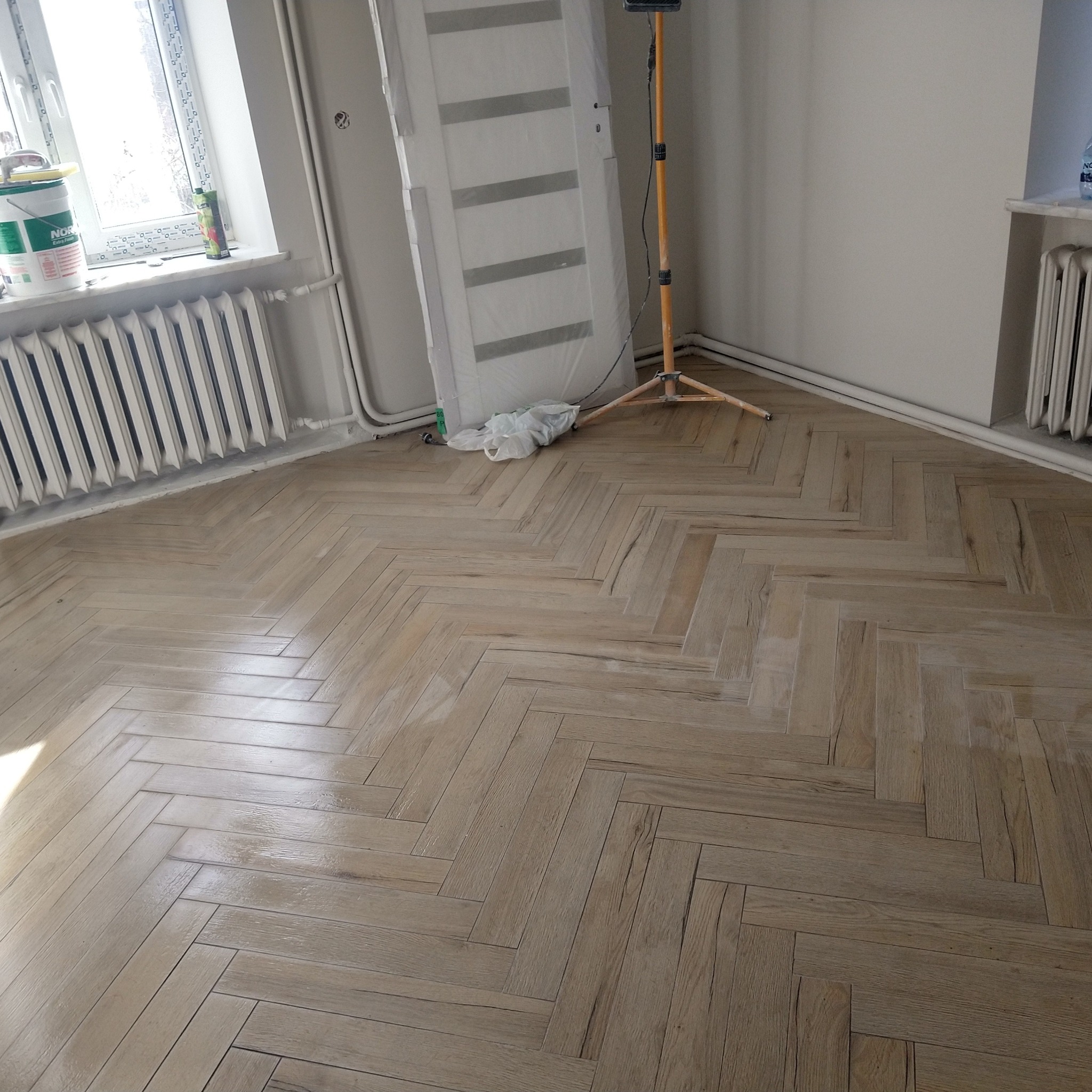 Ukończona podłoga z paneli ułożonych w jodełkę, widok z góry. W tle okno, grzejnik, drzwi w folii i lampa budowlana. Jasne wnętrze po remoncie.