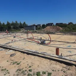 Przygotowany fundament pod budowę domu z instalacjami. Widoczne przewody i rury. Na drugim planie domy i zieleń.