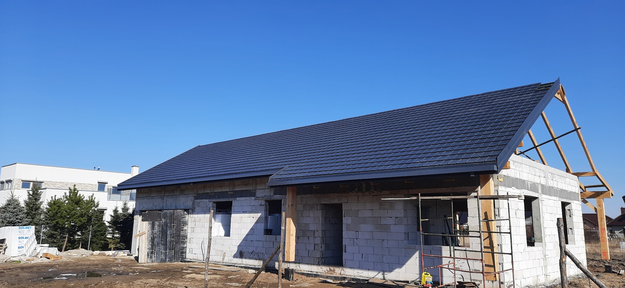 Budowa domu jednorodzinnego w Kruszwicy: ściany z betonu komórkowego, czarny dach z blachodachówki, widoczne rusztowania i elementy konstrukcji więźby dachowej.