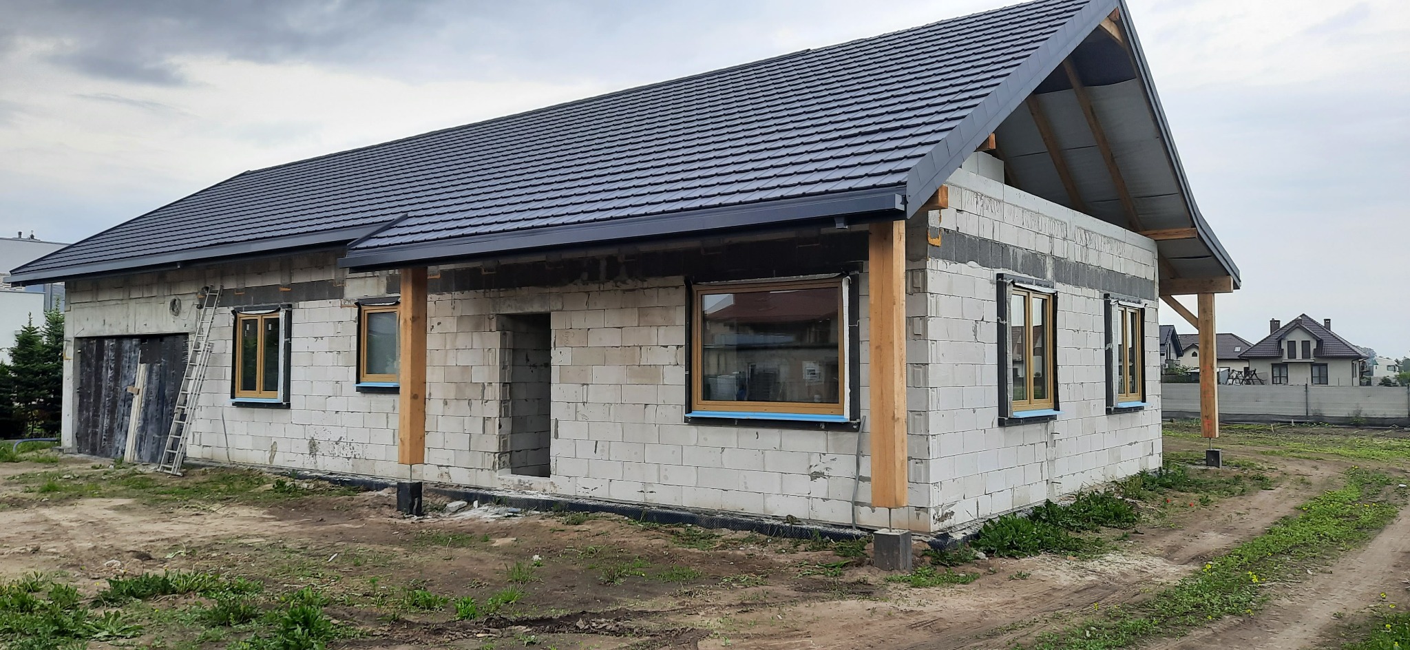 Surowy dom jednorodzinny z szarej cegły, z zamontowanymi oknami w drewnianych ramach i czarnym dachem z dachówki. Na ścianie oparta drabina.