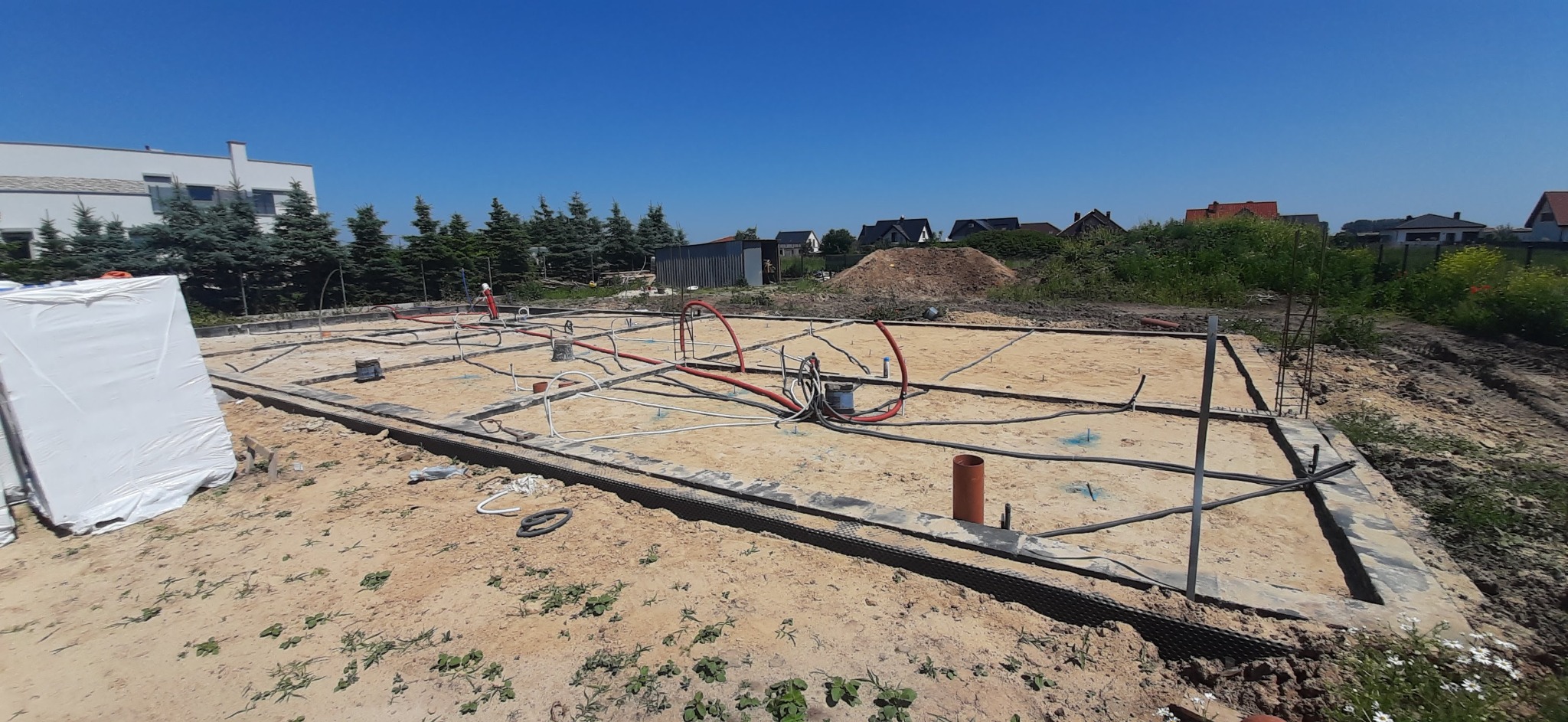 Przygotowany fundament pod budowę domu z instalacjami. Widoczne przewody i rury. Na drugim planie domy i zieleń.