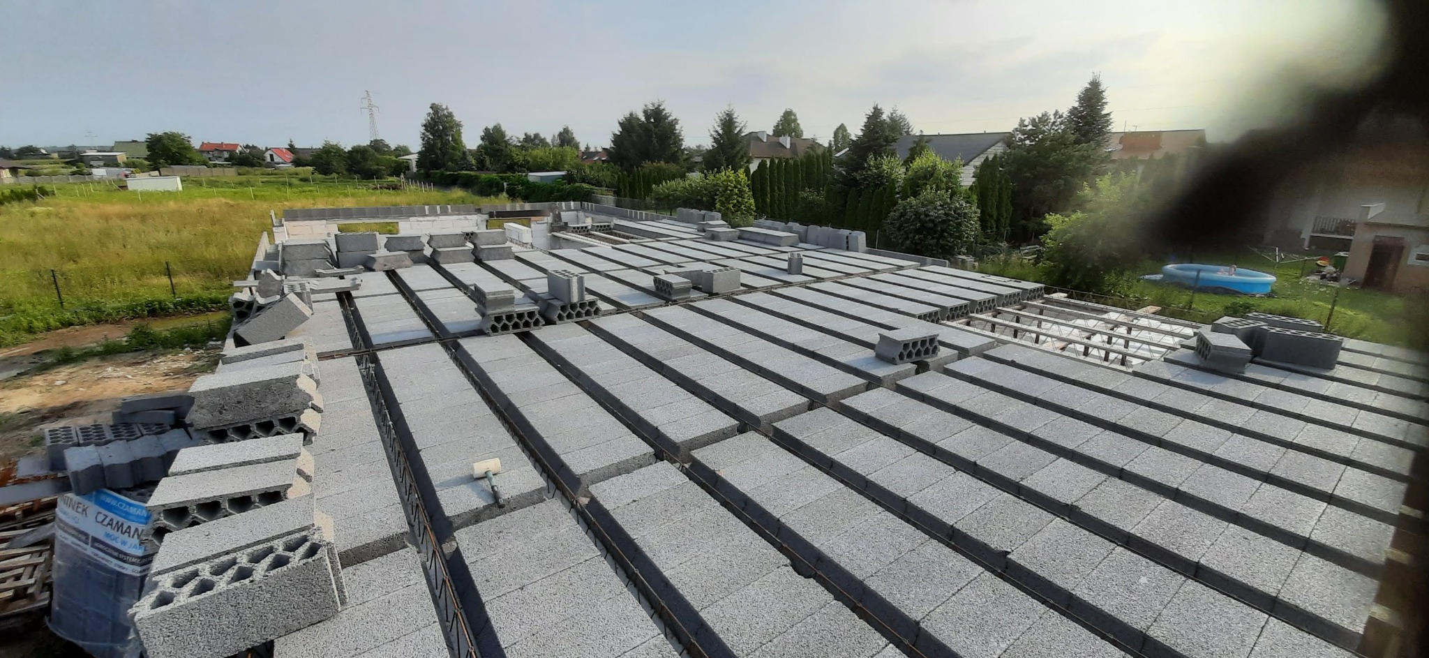 Budowa stropu Teriva: pustaki ułożone na belkach, widoczne zbrojenie. W tle zielone drzewa i zabudowa jednorodzinna. Etap prac przed zalaniem betonem.