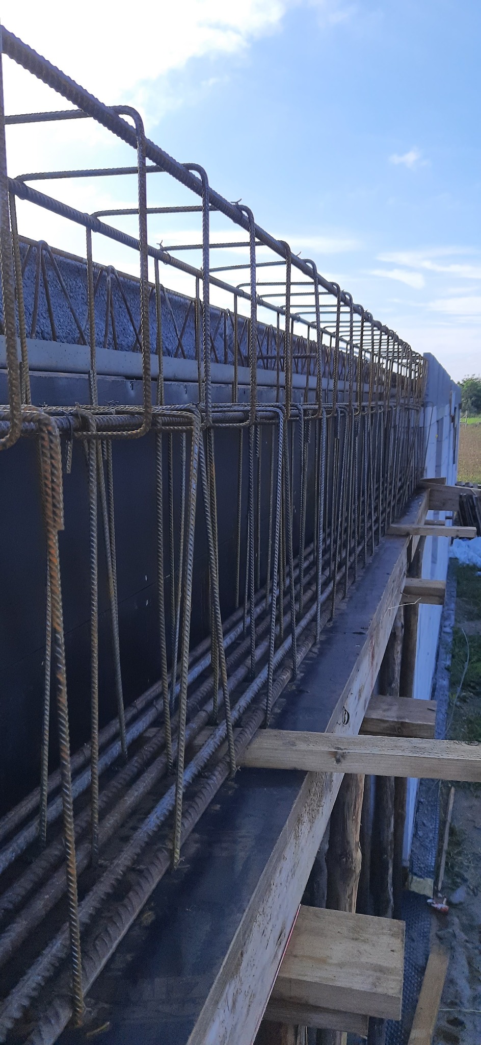 Zbrojenie fundamentów z widocznymi prętami stalowymi w szalunku, przygotowane do zalania betonem. Widok z góry, niebo w tle.