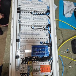 AMPERVOJT Wojciech Łakomski - Rozdzielnica elektryczna w białej obudowie z licznymi bezpiecznikami, przewodami w kolorach niebieskim, żółtym i czarnym. Widoczne elementy marki Eaton, Hager i Simtec.