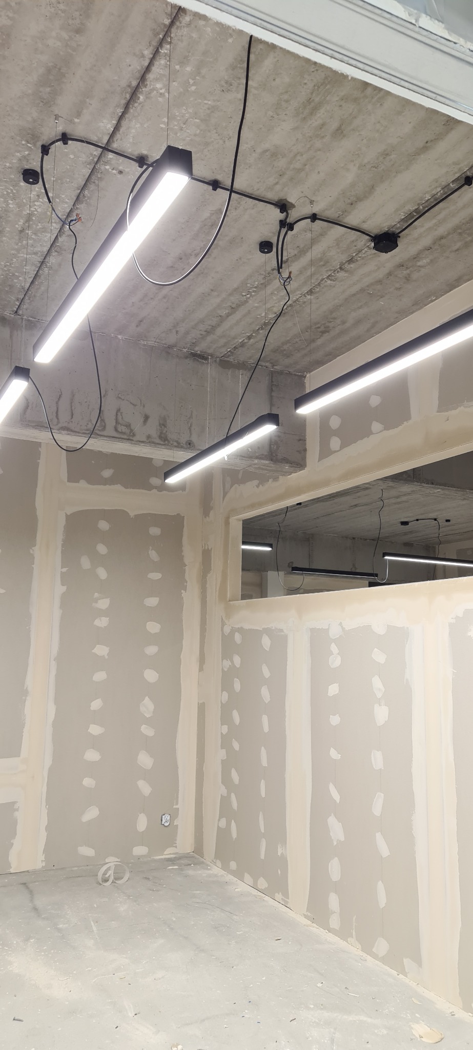 Montaż podwieszanych lamp LED z czarnymi przewodami na surowym, betonowym suficie w lokalu z płytami gipsowo-kartonowymi przed malowaniem. Widok z dołu.