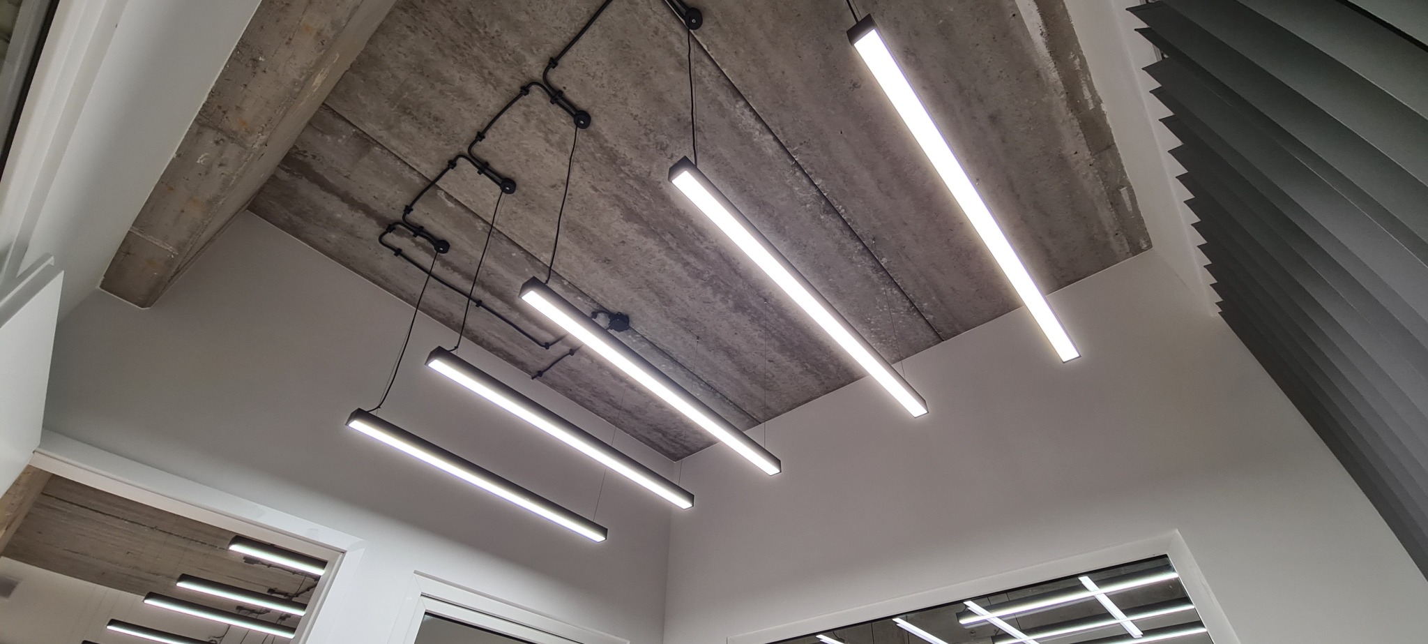 Nowoczesne, podwieszone oświetlenie LED w biurze z surowym, betonowym stropem i widocznymi przewodami. Minimalistyczny design lamp.