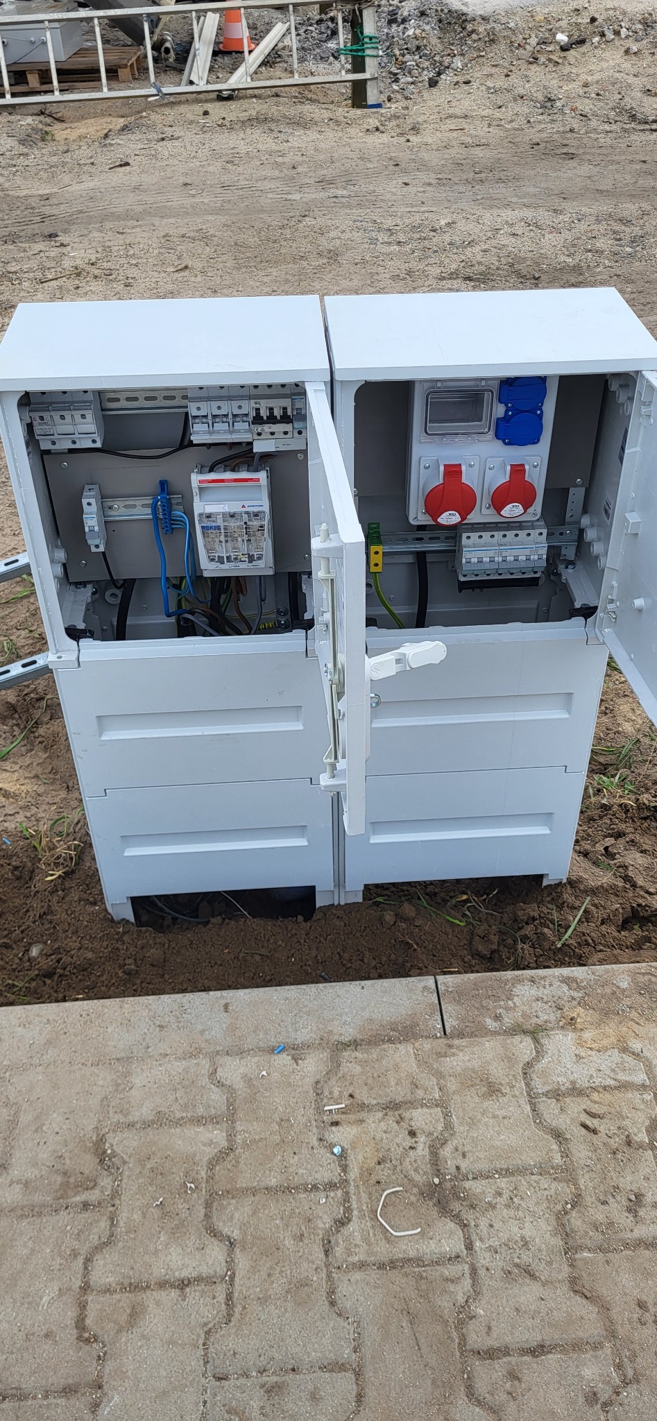 Otwarta, biała skrzynka elektryczna z widocznymi bezpiecznikami, gniazdami i okablowaniem, umieszczona w ziemi obok chodnika z kostki brukowej. Widoczne drabina i pachołek w tle.