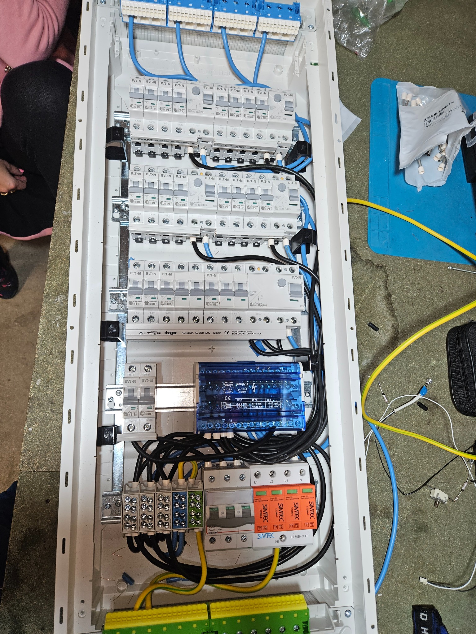 Rozdzielnica elektryczna w białej obudowie z licznymi bezpiecznikami, przewodami w kolorach niebieskim, żółtym i czarnym. Widoczne elementy marki Eaton, Hager i Simtec.