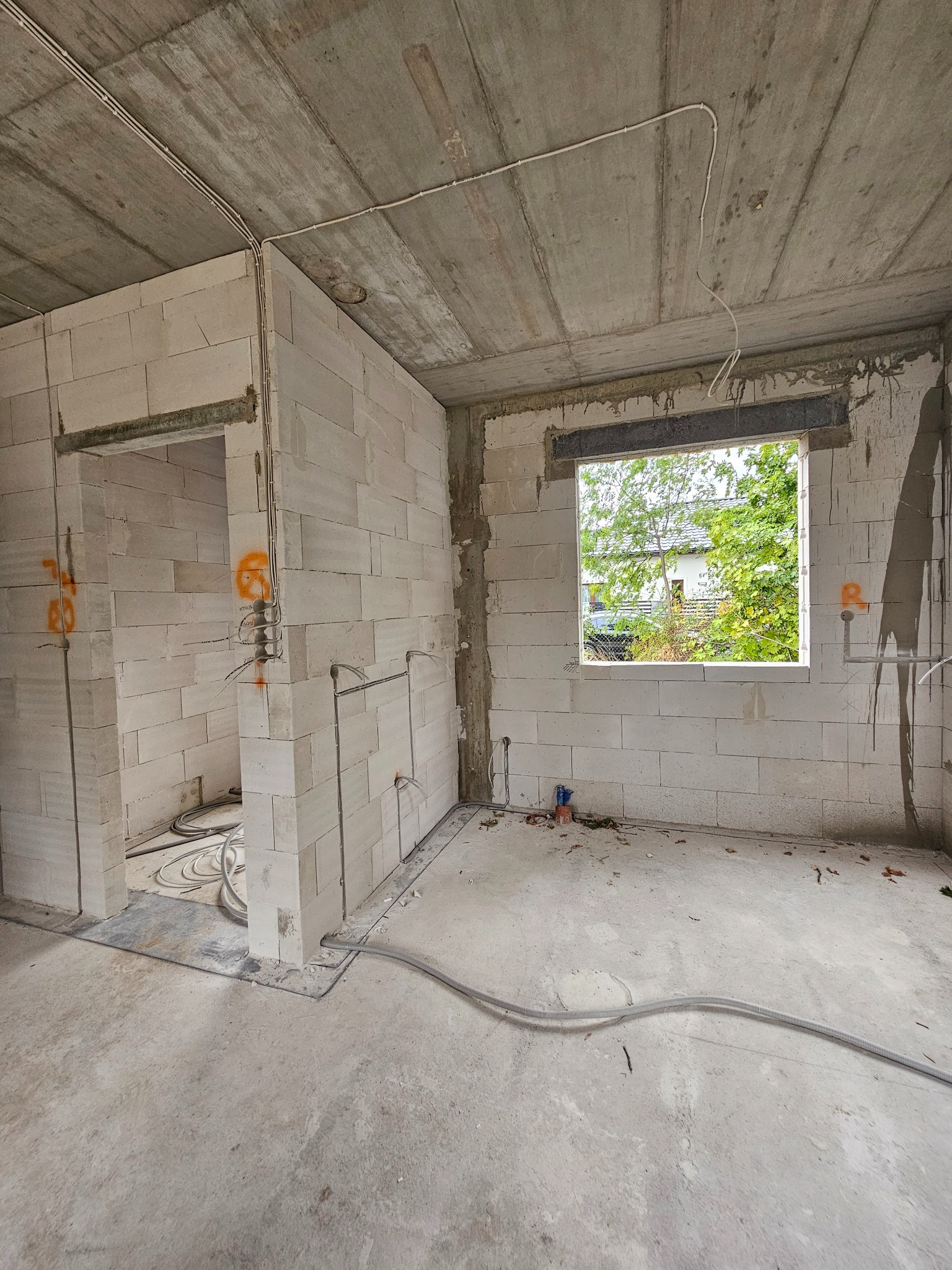 Wnętrze w stanie surowym z widoczną instalacją elektryczną w peszlu, prowadzoną po ścianach z betonu komórkowego i pod sufitem. Widoczne okno z zielenią na zewnątrz.