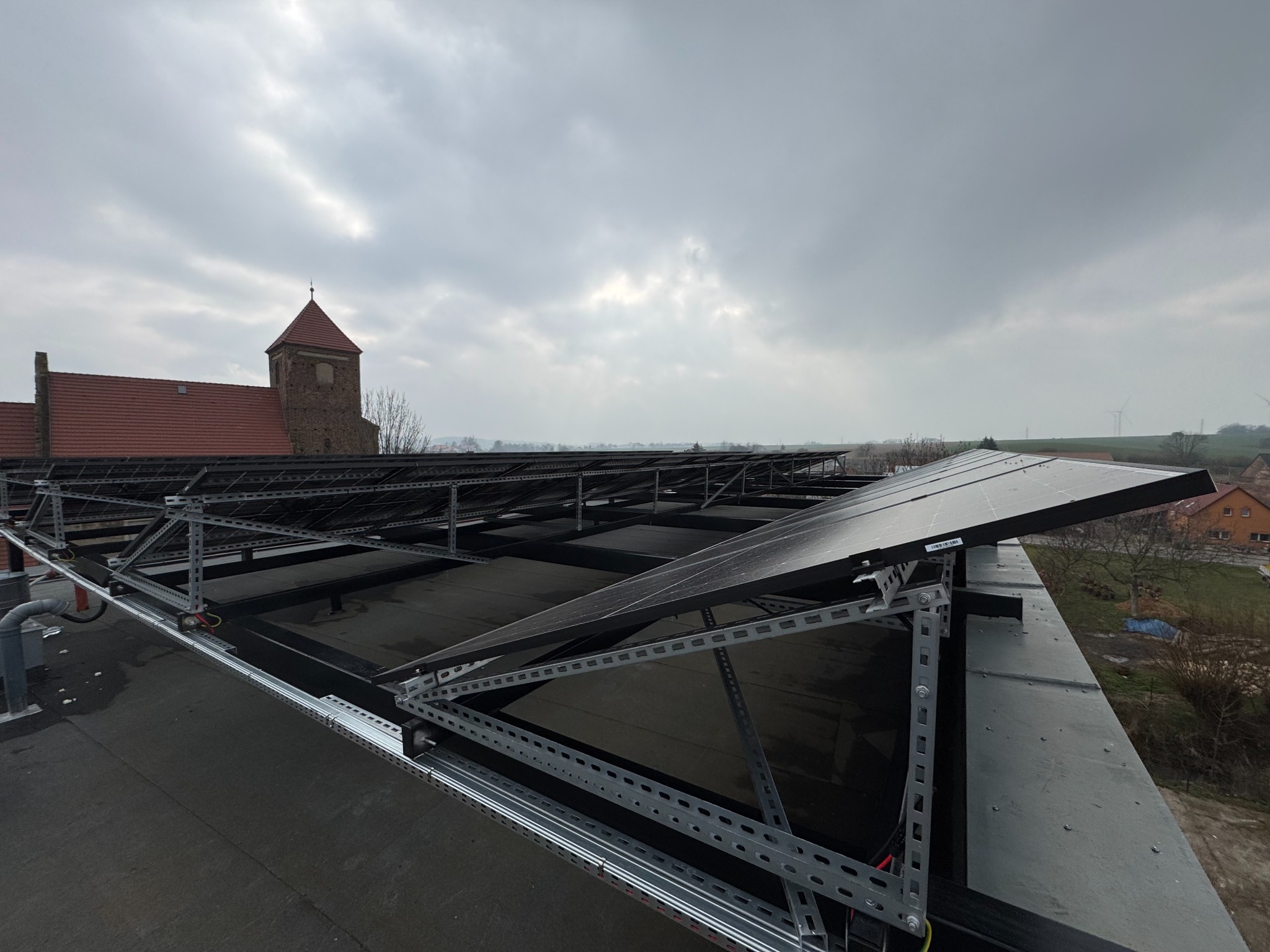 Montaż paneli słonecznych na dachu budynku z widoczną konstrukcją wsporczą i elementami instalacji elektrycznej. W tle kościół i pochmurne niebo.