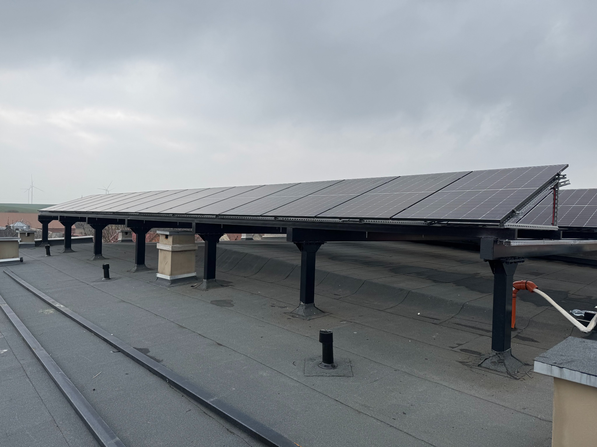 Instalacja paneli słonecznych na dachu budynku z widocznymi turbinami wiatrowymi w tle, pochmurne niebo, ujęcie z góry, czarne podpory konstrukcji.