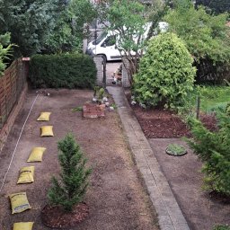 MKKA - Widok z góry na ogród w trakcie prac: świeżo posiana trawa, małe drzewko z korą, worki z ziemią w rzędzie, ścieżka z kostki, taczka i częściowo ułożona cegła.