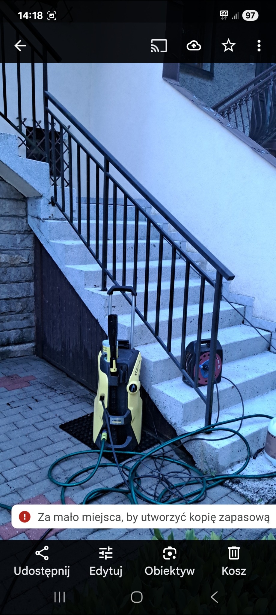 Żółto-czarna myjka ciśnieniowa Karcher na tle schodów zewnętrznych z czarną balustradą. Widoczny zwinięty wąż i kostka brukowa. Sprzątanie po umyciu.