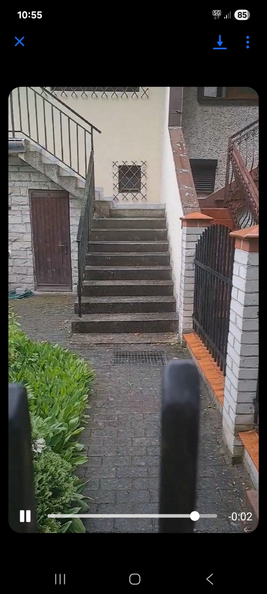 Schody wejściowe do budynku z kostki brukowej, balustrady po bokach, zielona roślinność po lewej stronie. Widok z dołu, częściowo zasłonięty przez element ogrodzenia.