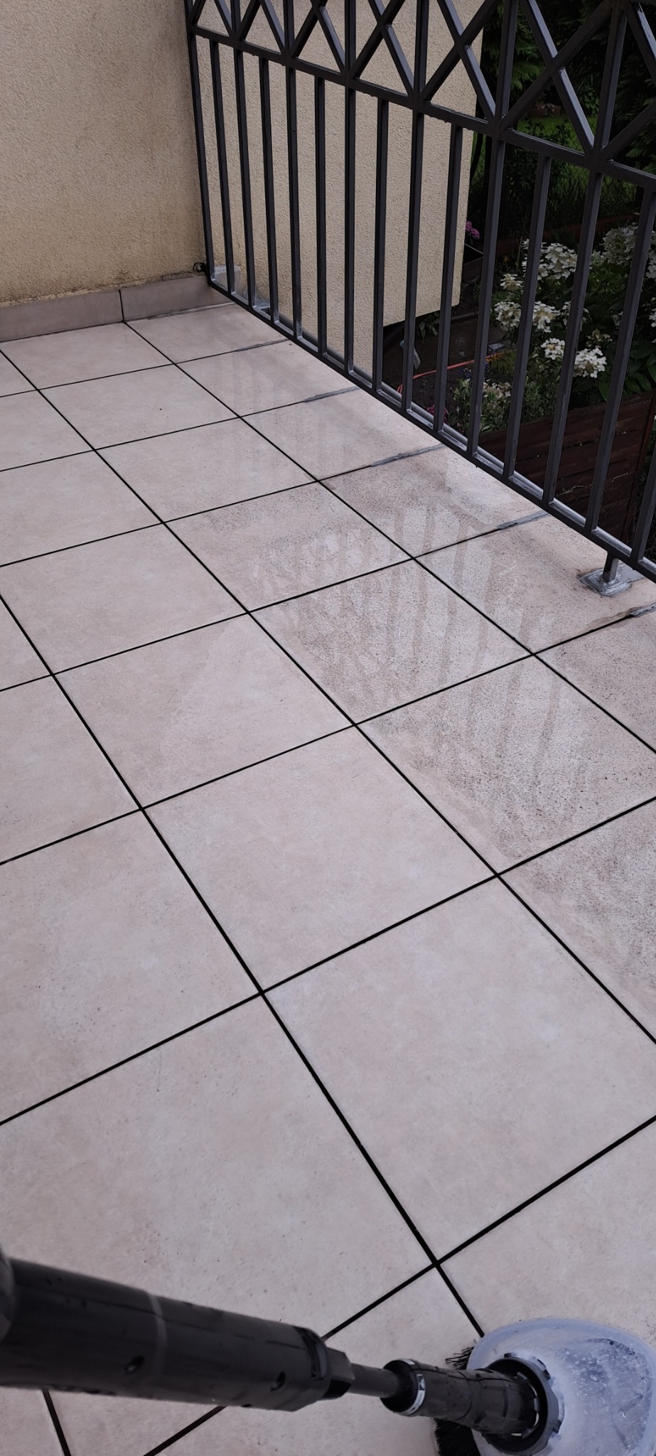 Czyszczenie płytek na balkonie za pomocą szczotki obrotowej pod ciśnieniem. Widoczne różnice w czystości płytek, balustrada i roślinność w tle.