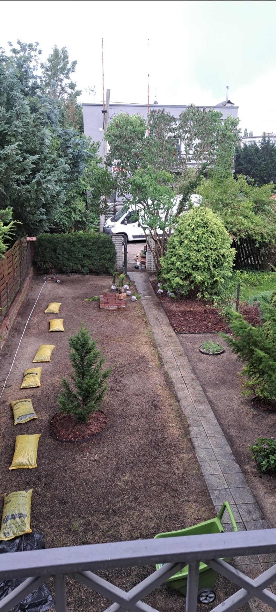 Widok z góry na ogród w trakcie prac: świeżo posiana trawa, małe drzewko z korą, worki z ziemią w rzędzie, ścieżka z kostki, taczka i częściowo ułożona cegła.