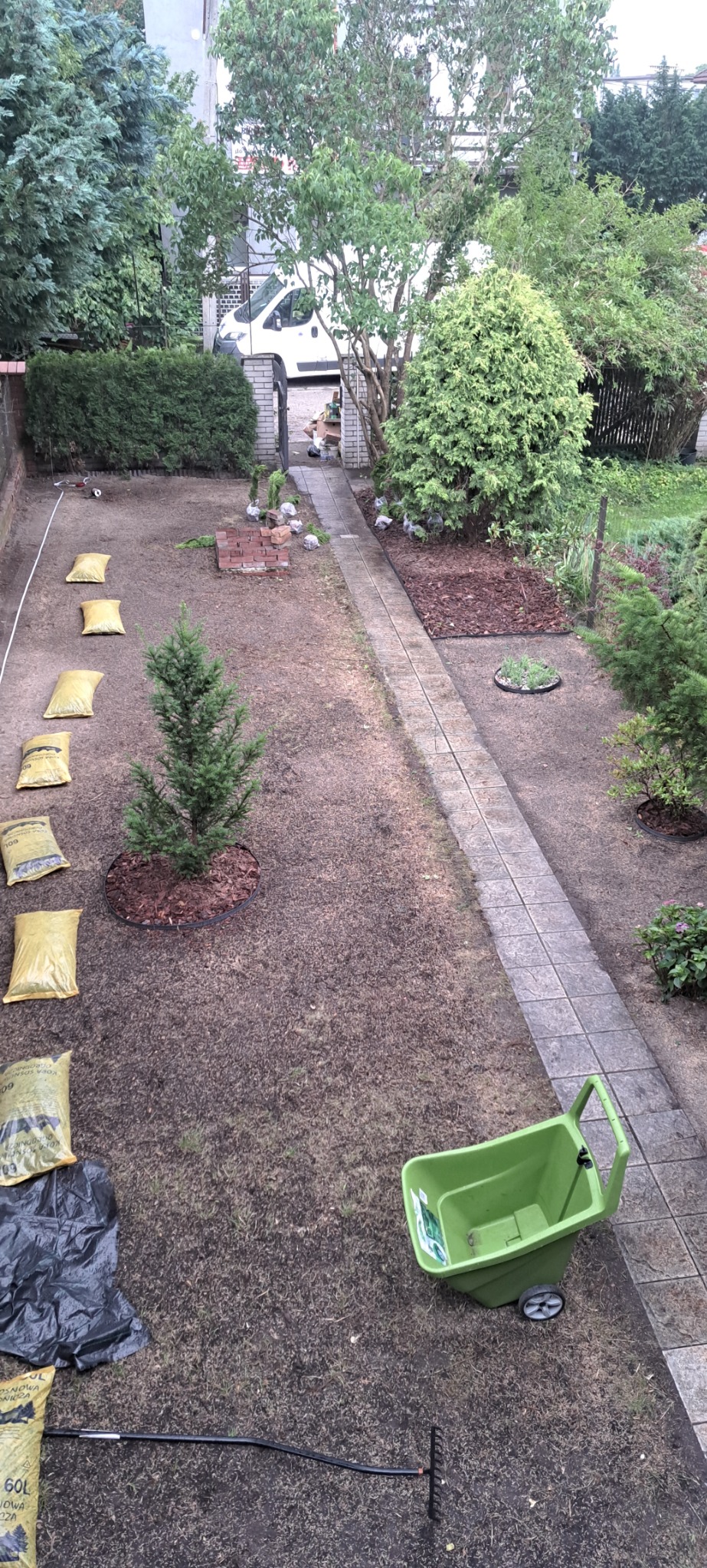 Przygotowany teren pod trawnik: worki z ziemią, sadzonki drzewek, zielony wózek ogrodowy i grabie. Widok z góry na działkę przed założeniem trawnika.