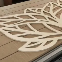 Wycinany laserowo, ażurowy dekor z jasnej sklejki na stole frezarki CNC. Wzór roślinny z powtarzalnymi elementami liści. Widoczne ślady frezowania.