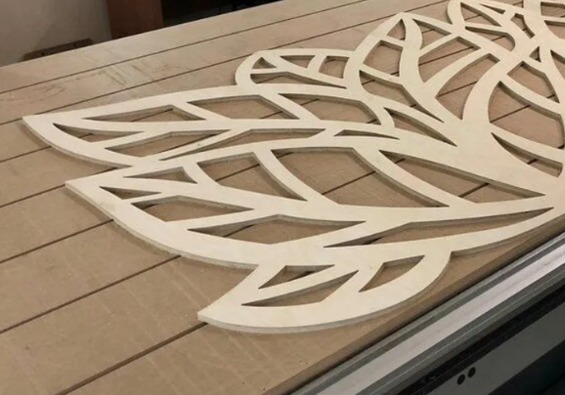 Wycinany laserowo, ażurowy dekor z jasnej sklejki na stole frezarki CNC. Wzór roślinny z powtarzalnymi elementami liści. Widoczne ślady frezowania.