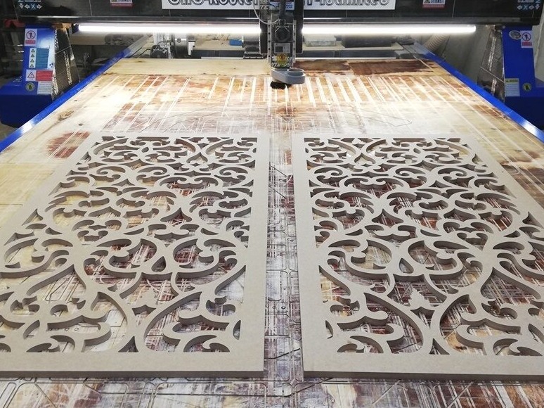 Dwa panele dekoracyjne z MDF z wyciętym ornamentem na stole frezarki CNC. Ujęcie z góry, widoczna głowica frezarki i ślady obróbki na stole.