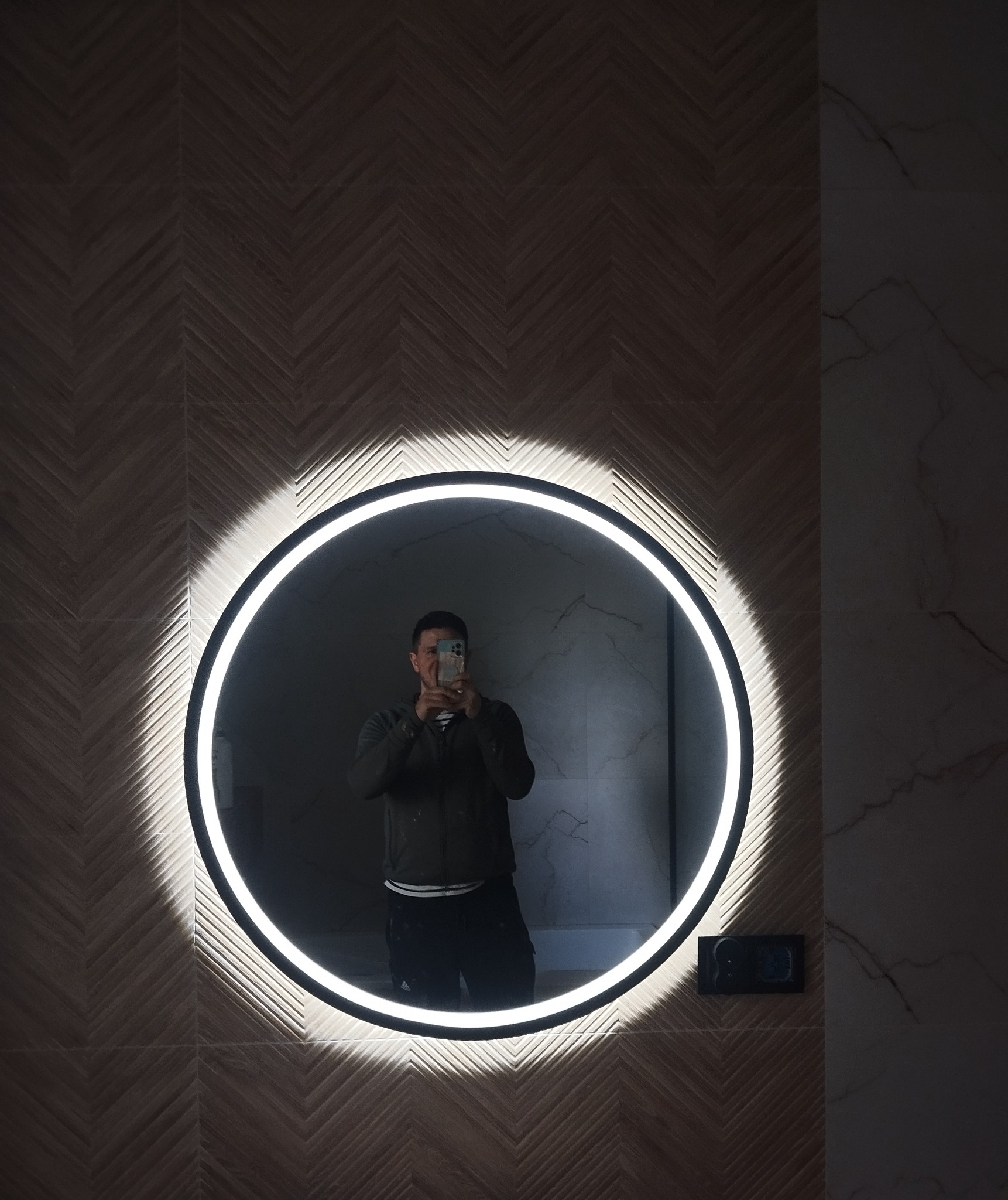 Okrągłe lustro z podświetleniem LED na ścianie z dekoracyjną płytką w jodełkę. Osoba robiąca zdjęcie widoczna w odbiciu.