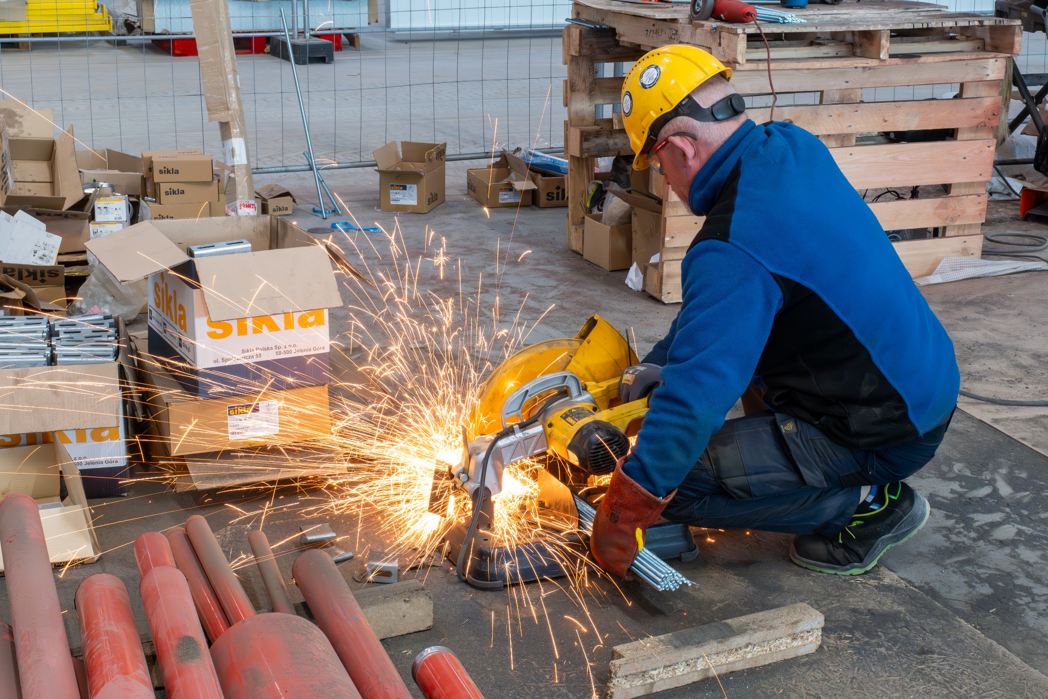 Pracownik w kasku tnie metalową rurę szlifierką kątową, iskry rozbryzgują się na boki. W tle kartony i paleta, co sugeruje miejsce pracy w budowie lub warsztacie.