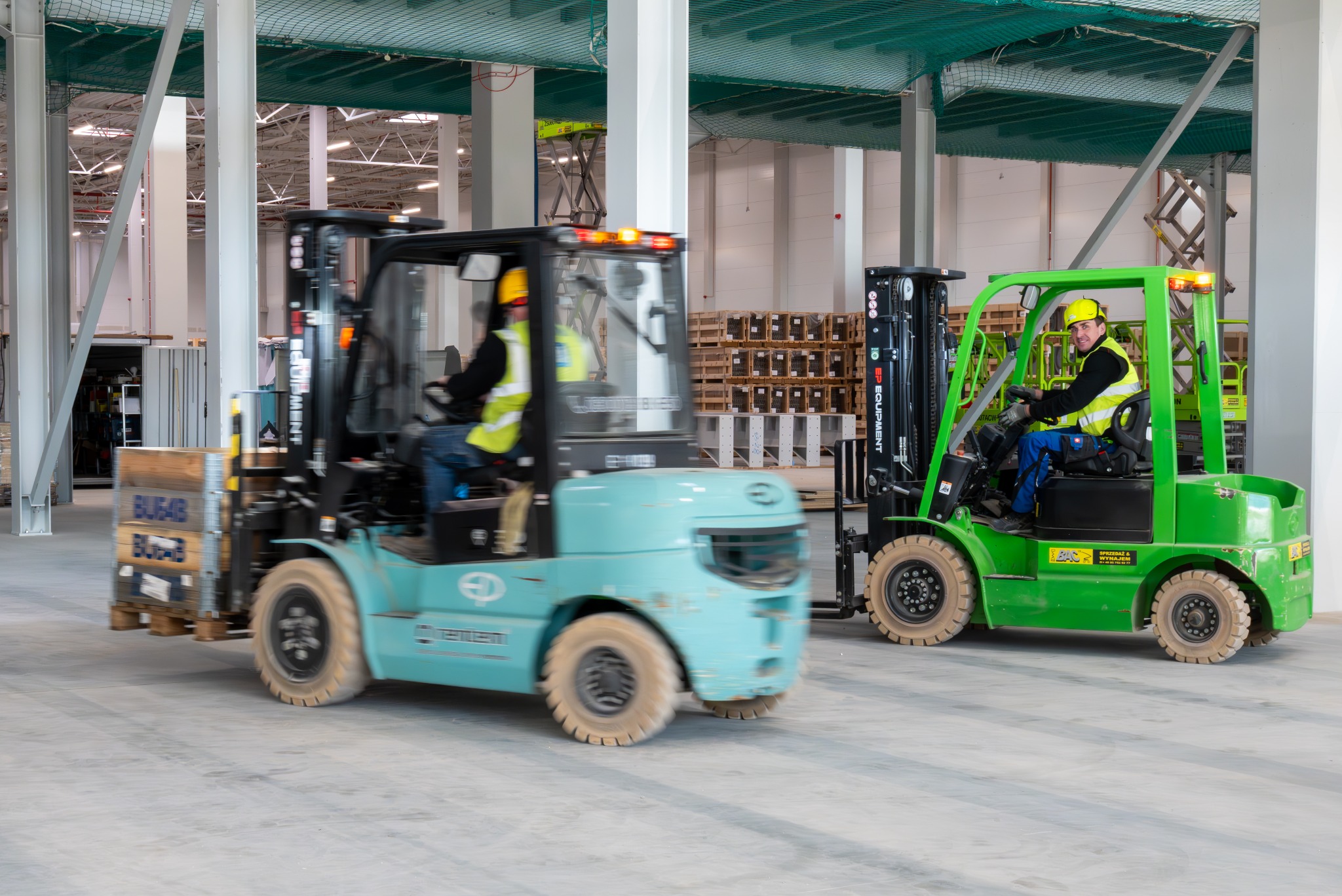 Dwa wózki widłowe w magazynie: turkusowy z ładunkiem i zielony z operatorem w kamizelce. Palety w tle, jasne oświetlenie hali, widoczna siatka zabezpieczająca pod sufitem.
