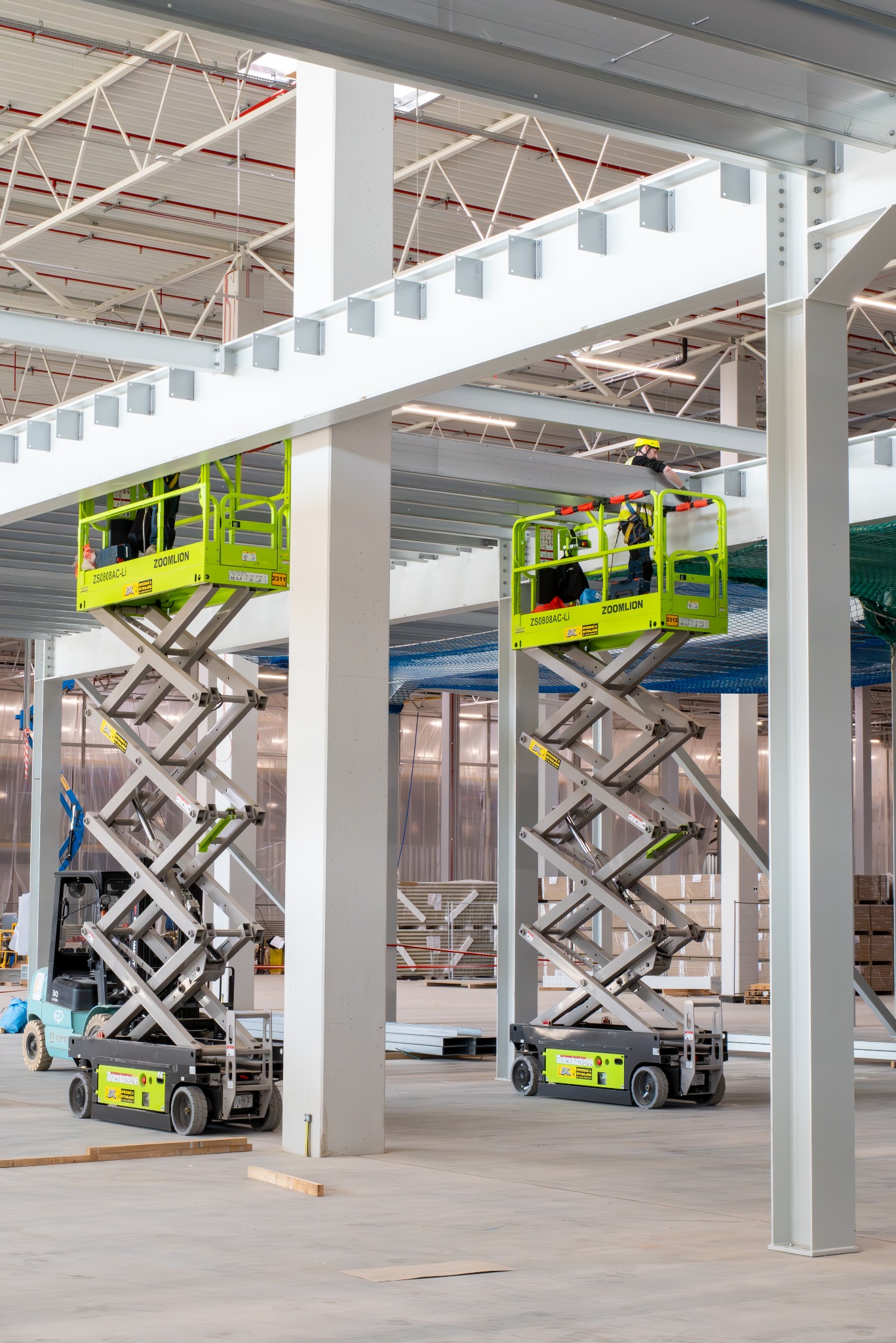 Montaż konstrukcji stalowej hali z użyciem podnośników nożycowych Zoomlion. Pracownicy w kaskach wykonują prace na wysokościach. Widoczny szkielet budynku i elementy konstrukcyjne.