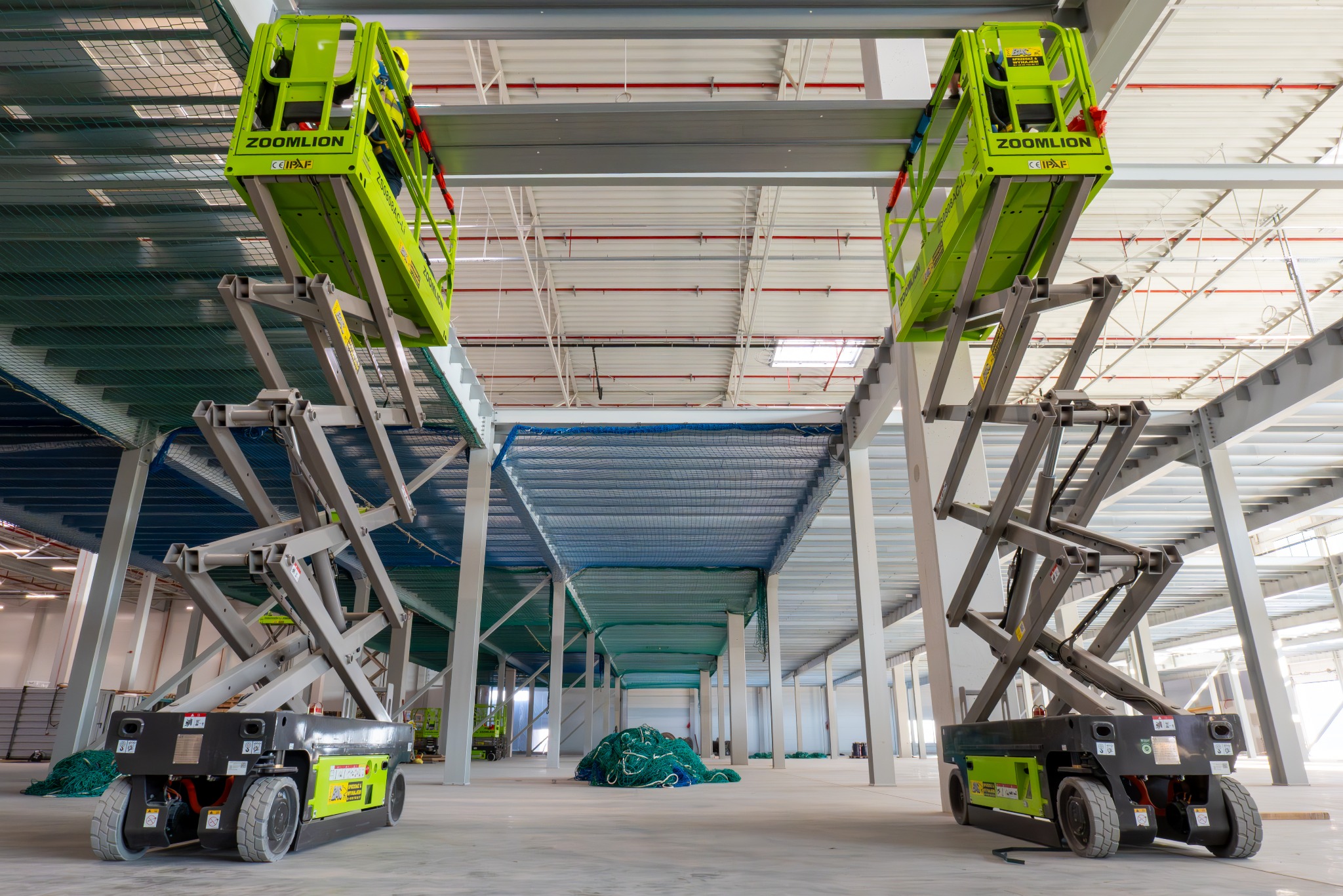 Wnętrze hali w trakcie budowy z dwoma podnośnikami nożycowymi Zoomlion, siatki zabezpieczające i stalowa konstrukcja dachu. Jasne oświetlenie.